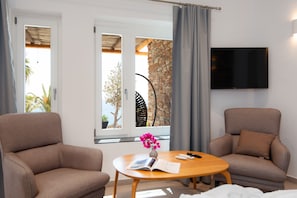 Quarto com dois sofás, uma mesa com um livro e controles remotos, televisão na parede e janela com vista para o mar e árvores.
