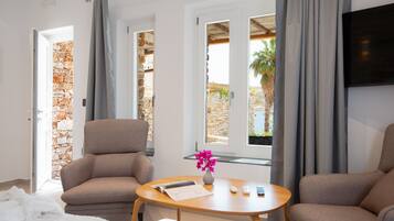 Une chambre d’hôtel avec un mur en pierre, un fauteuil beige, une table en bois ornée d’un vase de fleurs et une vue sur l’extérieur grâce à de grandes fenêtres.