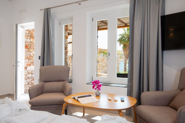 A hotel room with a stone wall, a beige armchair, a wooden table with a vase of flowers, and a view of the outdoors through large windows.
