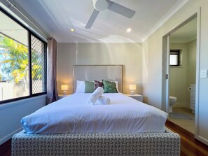 A hotel room with a large bed, a ceiling fan, a window with a view of palm trees, and a bathroom visible through an open door. - Waterfront Retreat with Pool & Pontoon, Broadbeach (Broadbeach Waters)
