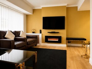 A living room with a brown leather sofa, a black coffee table, a flat-screen TV mounted on the wall, a fireplace, and a bench with a black cushion. - House Sleeps 4 With Parking (Sutton Coldfield)