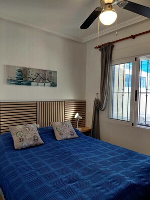 A bedroom with a bed, a nightstand, a lamp, a ceiling fan, a window with curtains, and a painting on the wall. - Bungalow ', La Veleta' with Mountain View, Shared Pool and Wi-Fi (Torrevieja)