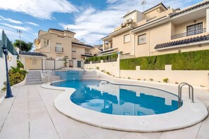 Ein Pool mit geschwungener Kante, umgeben von einem gepflasterten Weg, neben Wohngebäuden mit Balkonen und einer grünen Hecke.