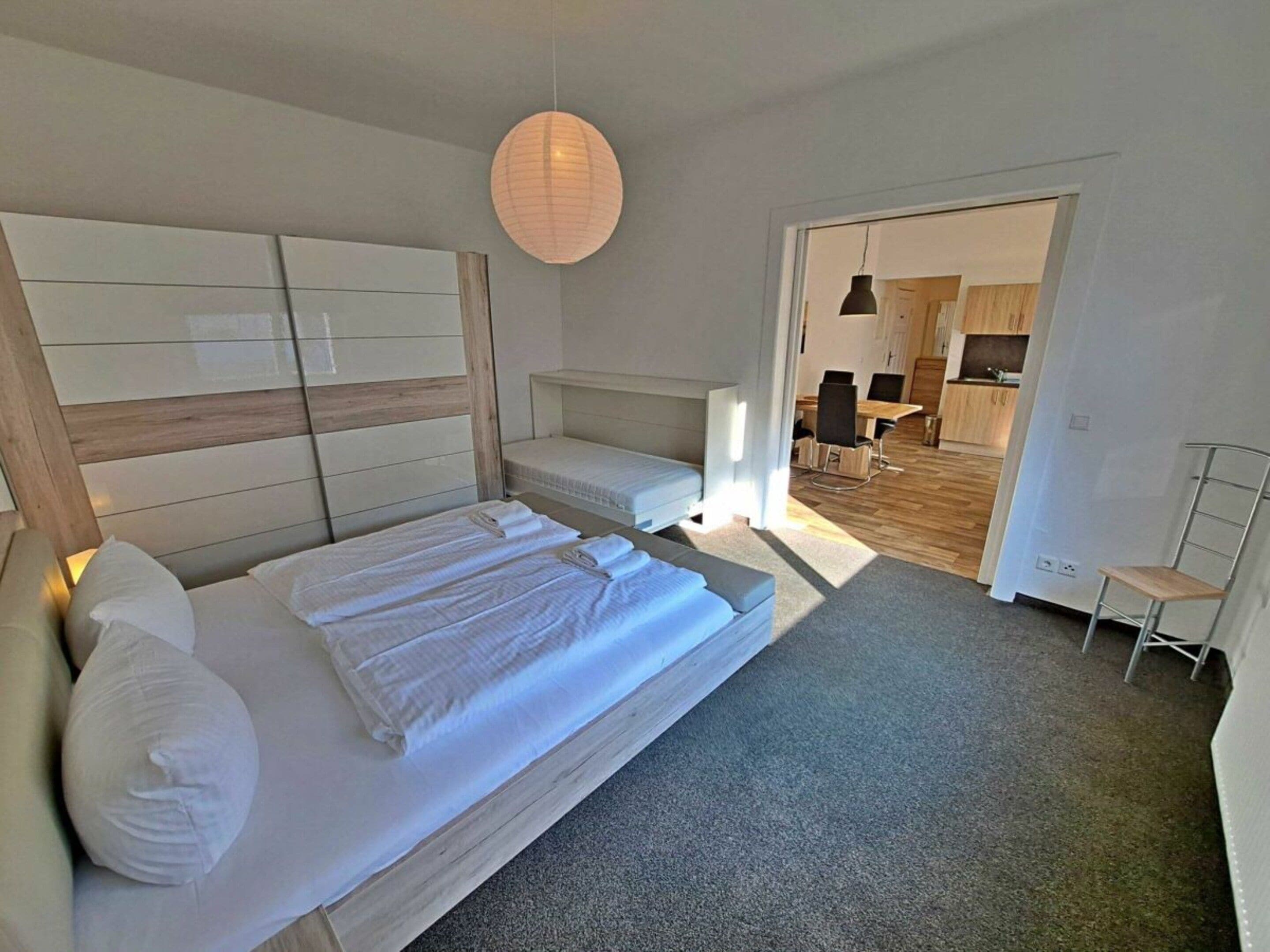 Une chambre avec un grand lit, une table de chevet, un luminaire suspendu et une vue sur la cuisine adjacente.