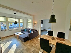 A modern living space with a sofa, dining table, and chairs, large windows with blinds, and a view of the outside. - Holiday Apartment With 1 Bedroom (Heringsdorf)