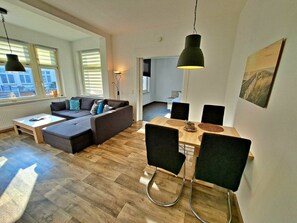 A modern living room with a sofa, dining table, and chairs. There are windows with blinds, a lamp, and a painting on the wall. - Holiday Apartment With 1 Bedroom (Heringsdorf)