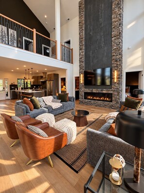A spacious living area with a fireplace, comfortable seating, and a kitchen in the background. - Luxury 9BR Massanutten Mountain Escape | Stunning Ski Slope Views, Hot Tub, Theater & Gym (McGaheysville)