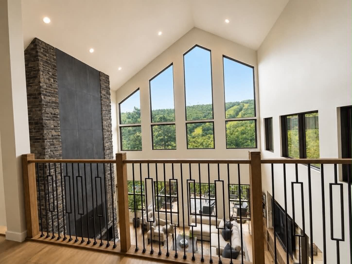 A spacious room with a high ceiling, large windows offering a view of greenery, and a stone fireplace.