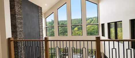A spacious room with a high ceiling, large windows offering a view of greenery, and a stone fireplace.