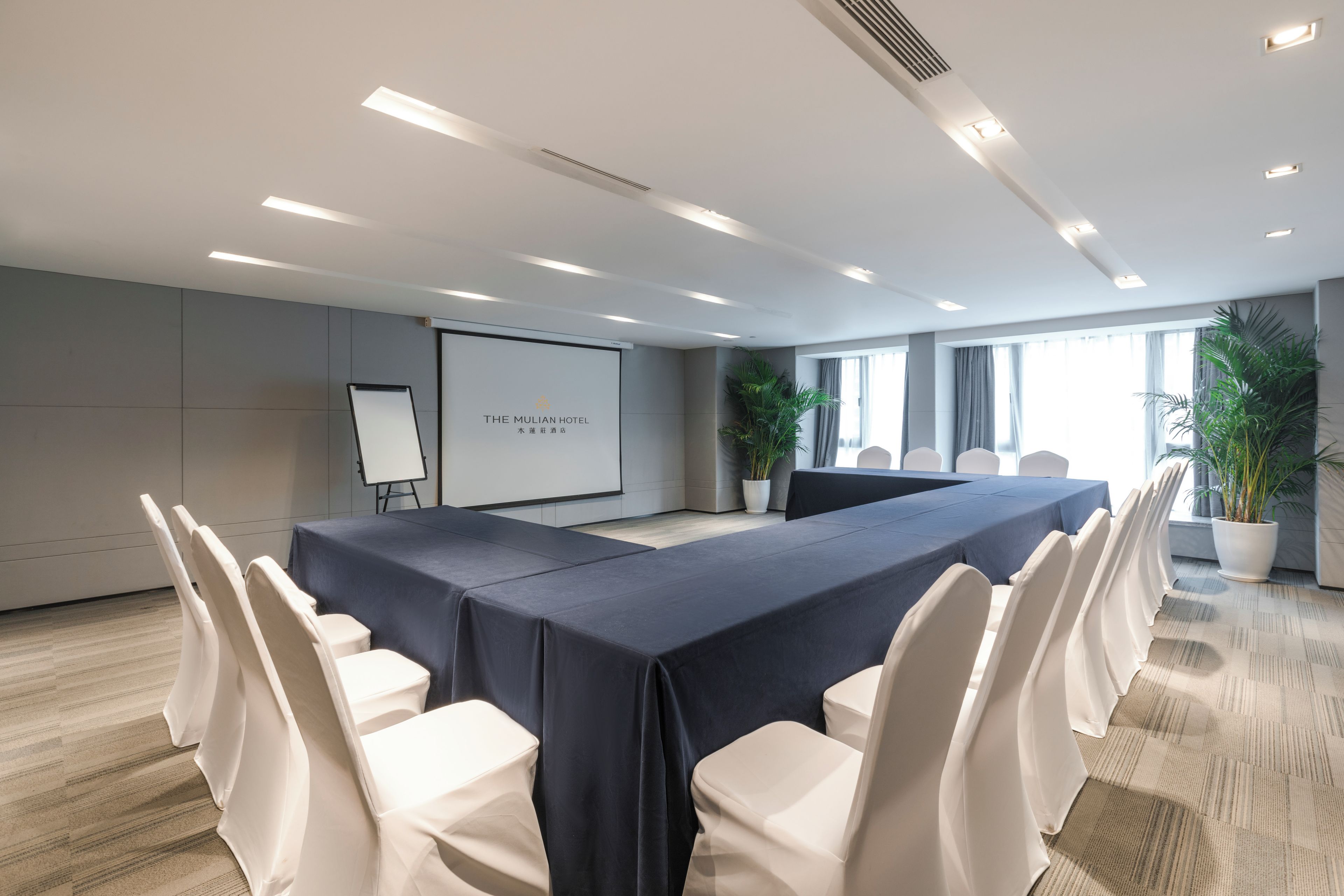 A conference room with a long table, white chairs, a projector screen, and potted plants.