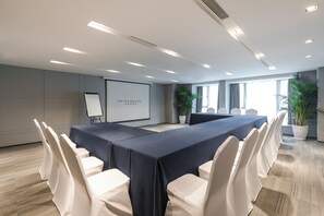 Un salón de conferencias con una larga mesa, sillas blancas, una pantalla de proyector y plantas en macetas.