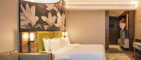 A hotel room with a bed, a sofa, a large floral wall art, a nightstand, and a bathroom visible in the background.