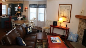 A living room with a brown leather sofa, a red coffee table, and a fireplace. There is a dining area with a table and chairs, and a kitchen with dark cabinets and a refrigerator.