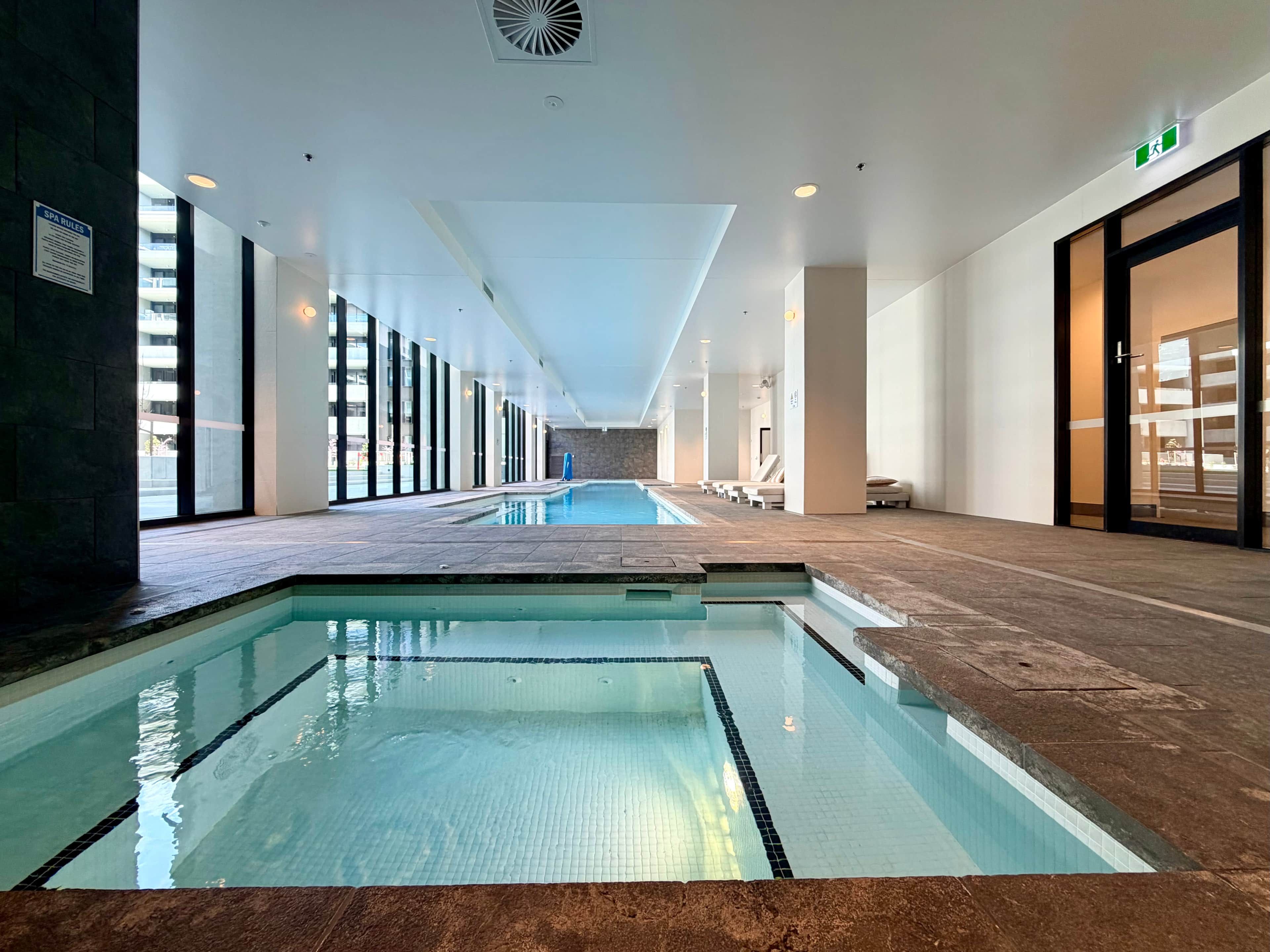 A modern indoor swimming pool with clear water, surrounded by a tiled floor and walls, and a long corridor with seating areas.