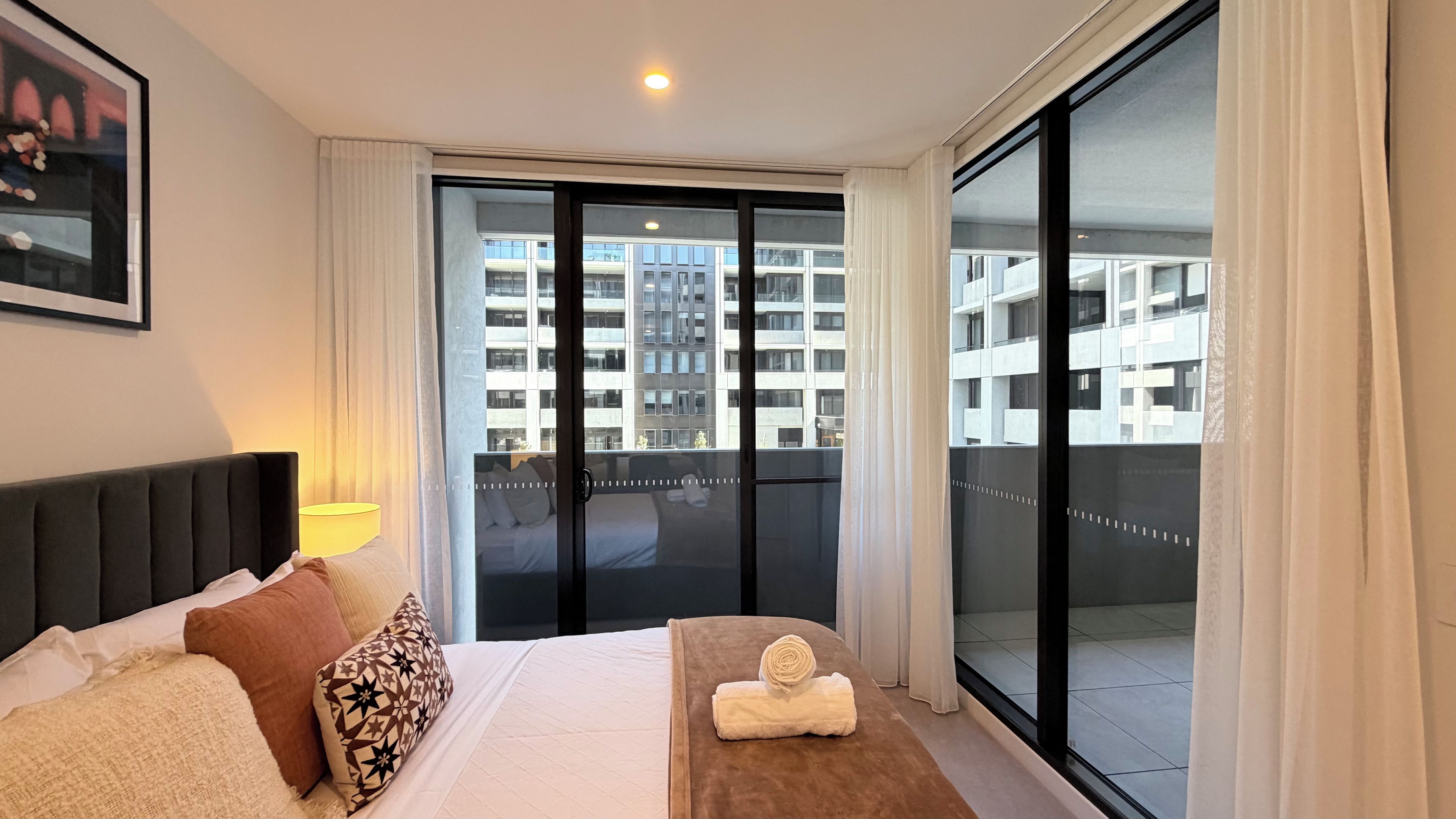 A hotel room with a bed, a bedside lamp, a window with a view of a building, and a small table with a towel.