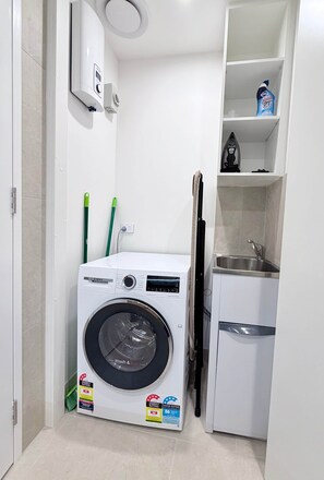 A compact laundry room with a Bosch washing machine, a small sink, and a shelf with cleaning supplies. - Elegant Parkes Stay near Lake & Parliament (Parkes)