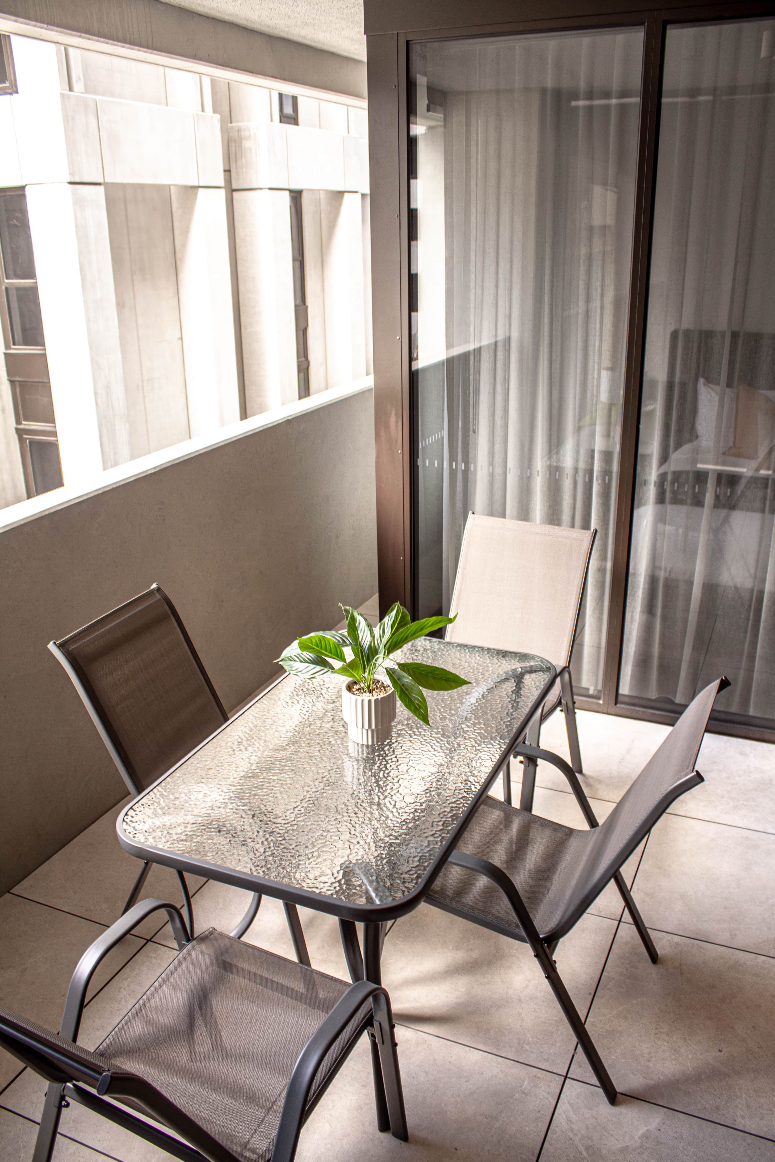 A balcony with a glass table, two chairs, a potted plant, and a view of buildings through the window.