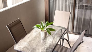 A balcony with a glass table, two chairs, a potted plant, and a view of buildings through the window.