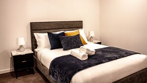 A neatly made bed with a dark headboard, white linens, and a variety of pillows. There are two bedside tables with lamps and a small drawer. - The Eastbourne Premium 2BR Central Canberra (Parkes)