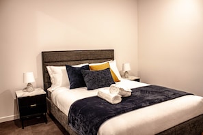 A neatly made bed with a dark headboard, white linens, and a variety of pillows. There are two bedside tables with lamps and a small drawer. - The Eastbourne Premium 2BR Central Canberra (Parkes)