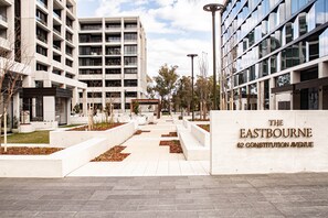 Um complexo moderno de edifícios com vários andares, um espaço aberto central com caminhos e uma placa indicando 'The Eastbourne 62 Constitution Avenue'.