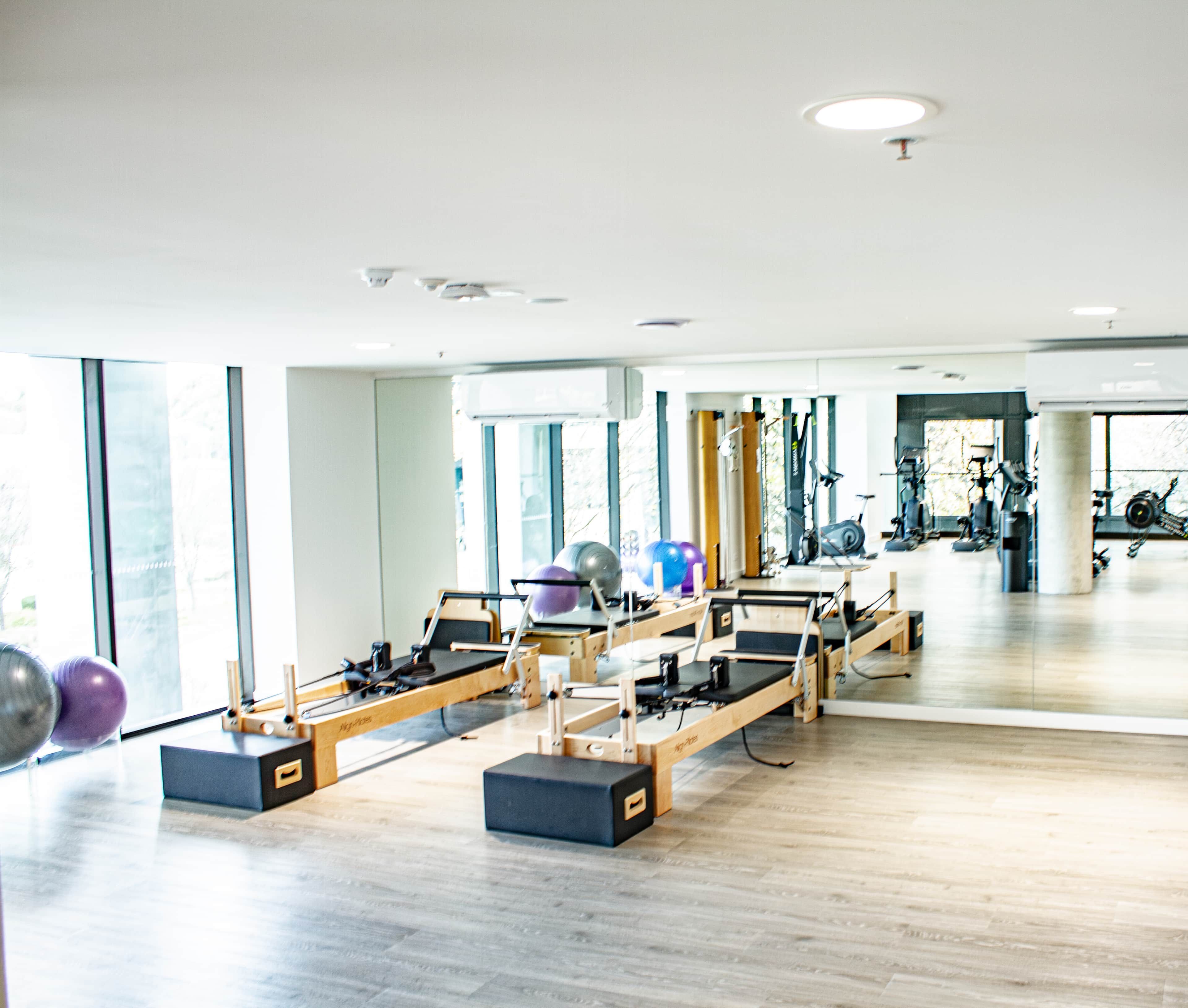 A Pilates studio with equipment, large windows, and a view of the gym area.