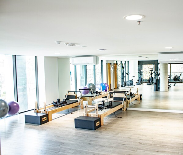 A Pilates studio with equipment, large windows, and a view of the gym area.