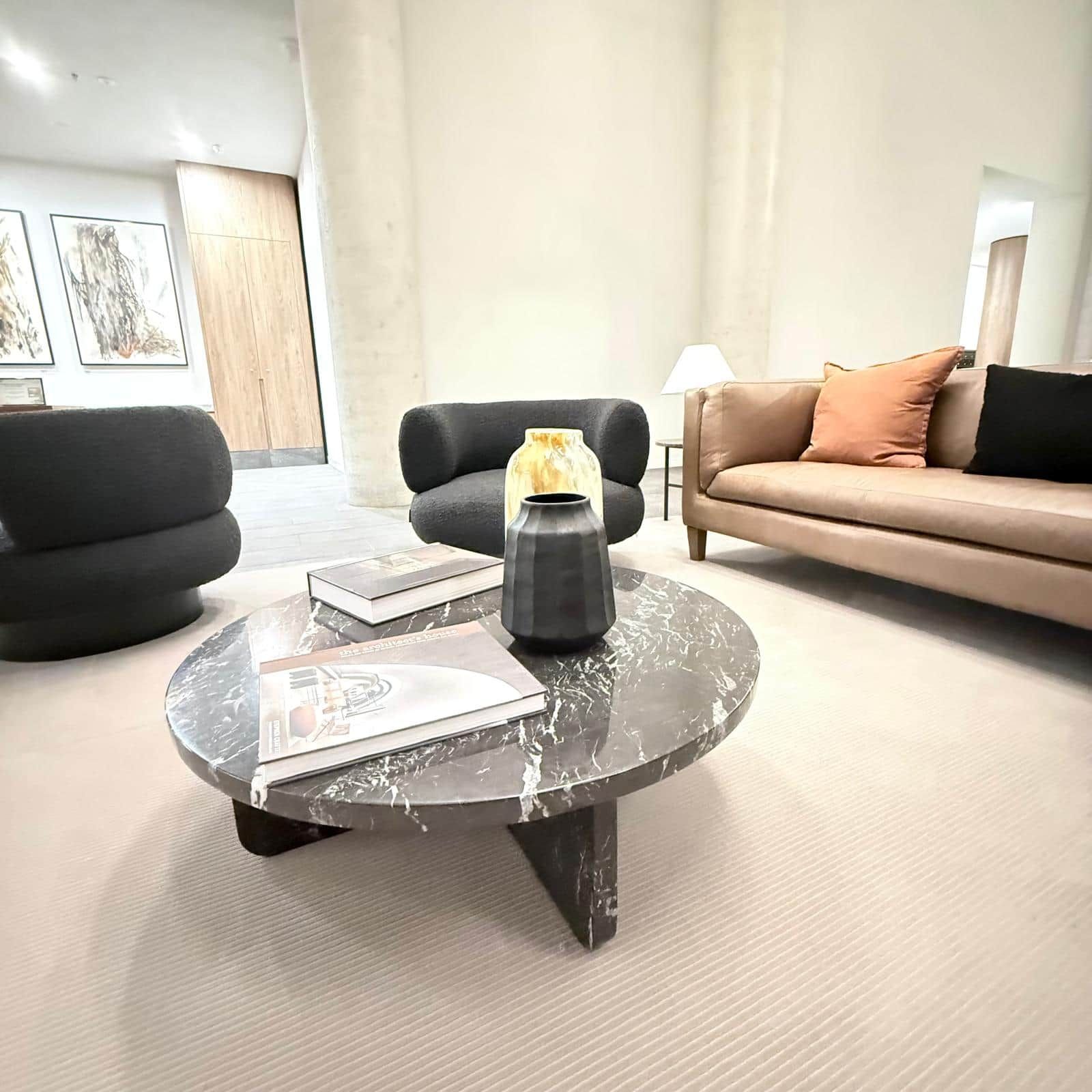 A modern living room with a round coffee table, two black armchairs, a beige sofa, and a lamp.