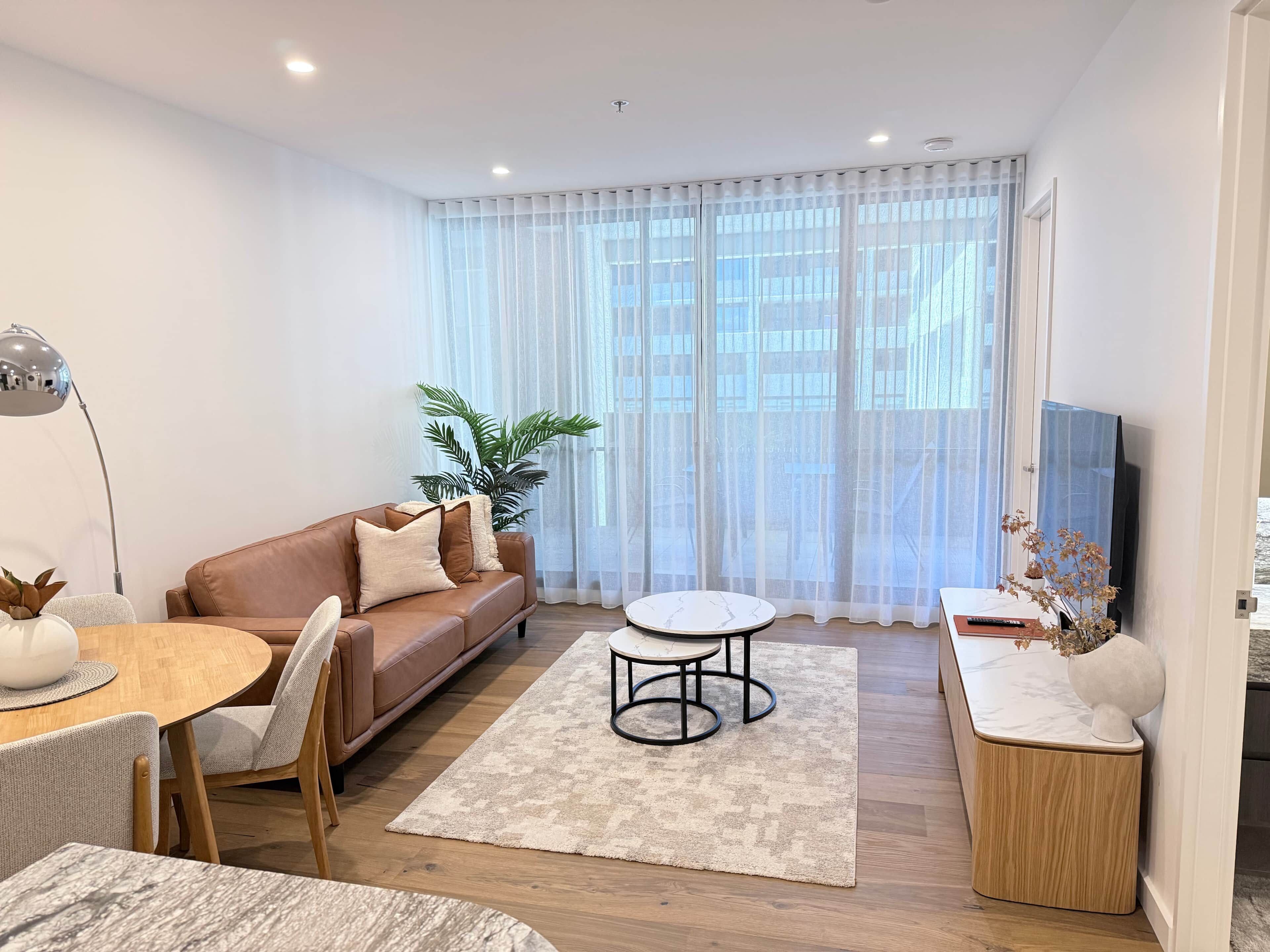 A modern living room with a brown leather sofa, a round dining table, and a TV on a stand.