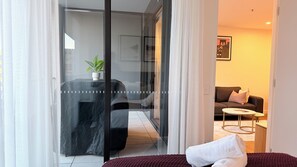 Et moderne hotelværelse med en glasdør, der fører til en balkon, en sofa, et lille rundt bord og en pænt redt seng med et håndklæde.