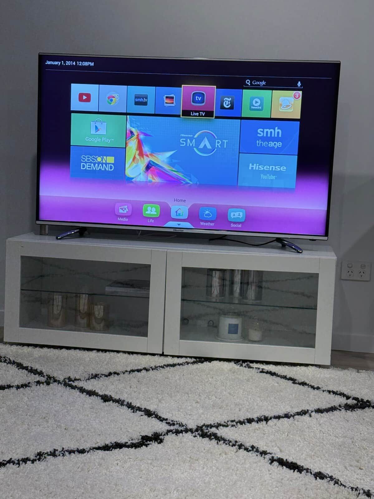 A flat-screen TV on a white cabinet with a glass front, displaying a home screen with various app icons and a date and time stamp.
