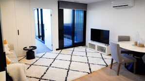 A modern living room with a flat-screen TV, a dining table with chairs, a patterned rug, and a sliding glass door leading to a balcony. - Stylish, Cosy City Retreat in Reid Canberra (Reid)
