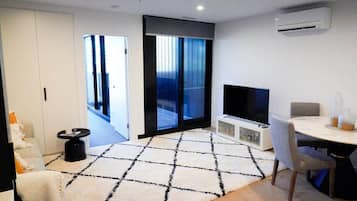 A modern living room with a flat-screen TV, a dining table with chairs, a patterned rug, and a sliding glass door leading to a balcony.