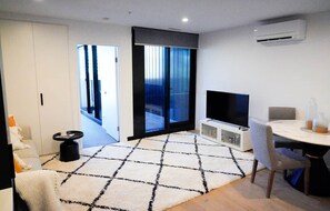 A modern living room with a flat-screen TV, a dining table with chairs, a patterned rug, and a sliding glass door leading to a balcony. - Stylish, Cosy City Retreat in Reid Canberra (Reid)