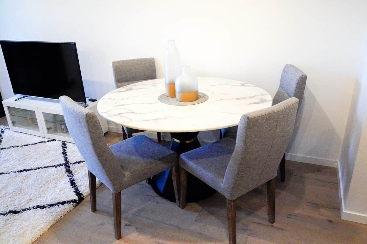 A dining area with a round table, four grey upholstered chairs, a white marble table base, and two glass bottles on the table.