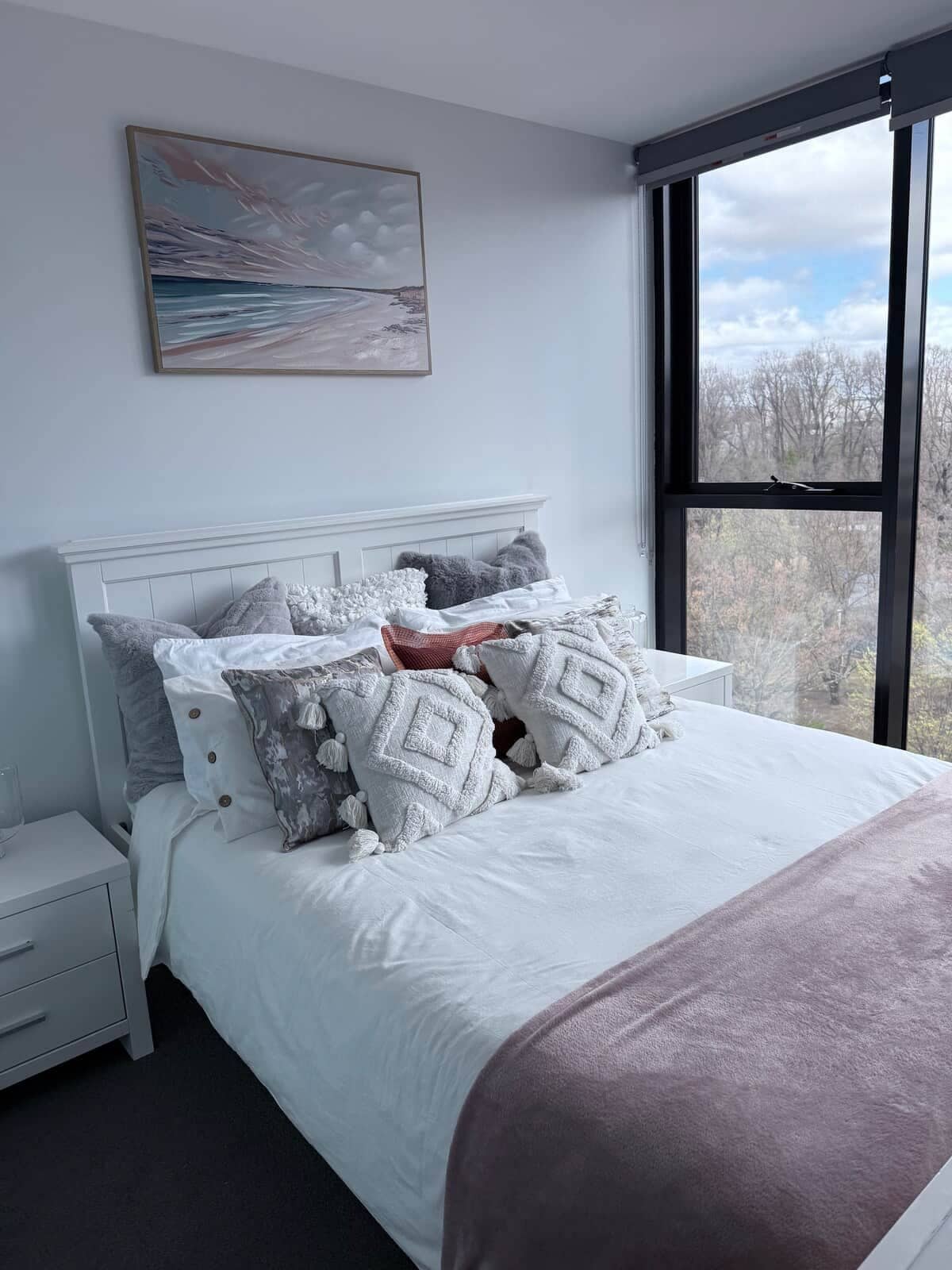 A bedroom with a large bed, a nightstand, a window with a view, and a painting on the wall.