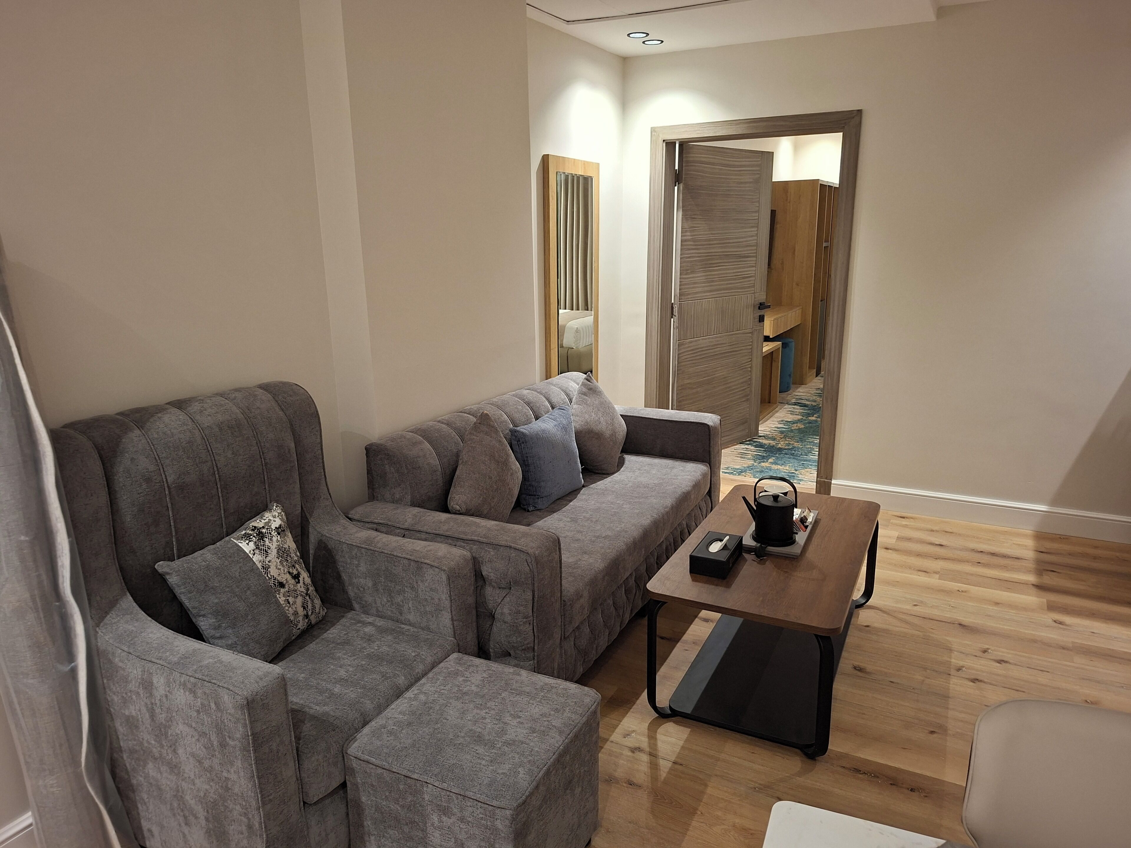 A modern living room with a grey sofa set, a small wooden coffee table, and a view into a bedroom with a wooden wardrobe.