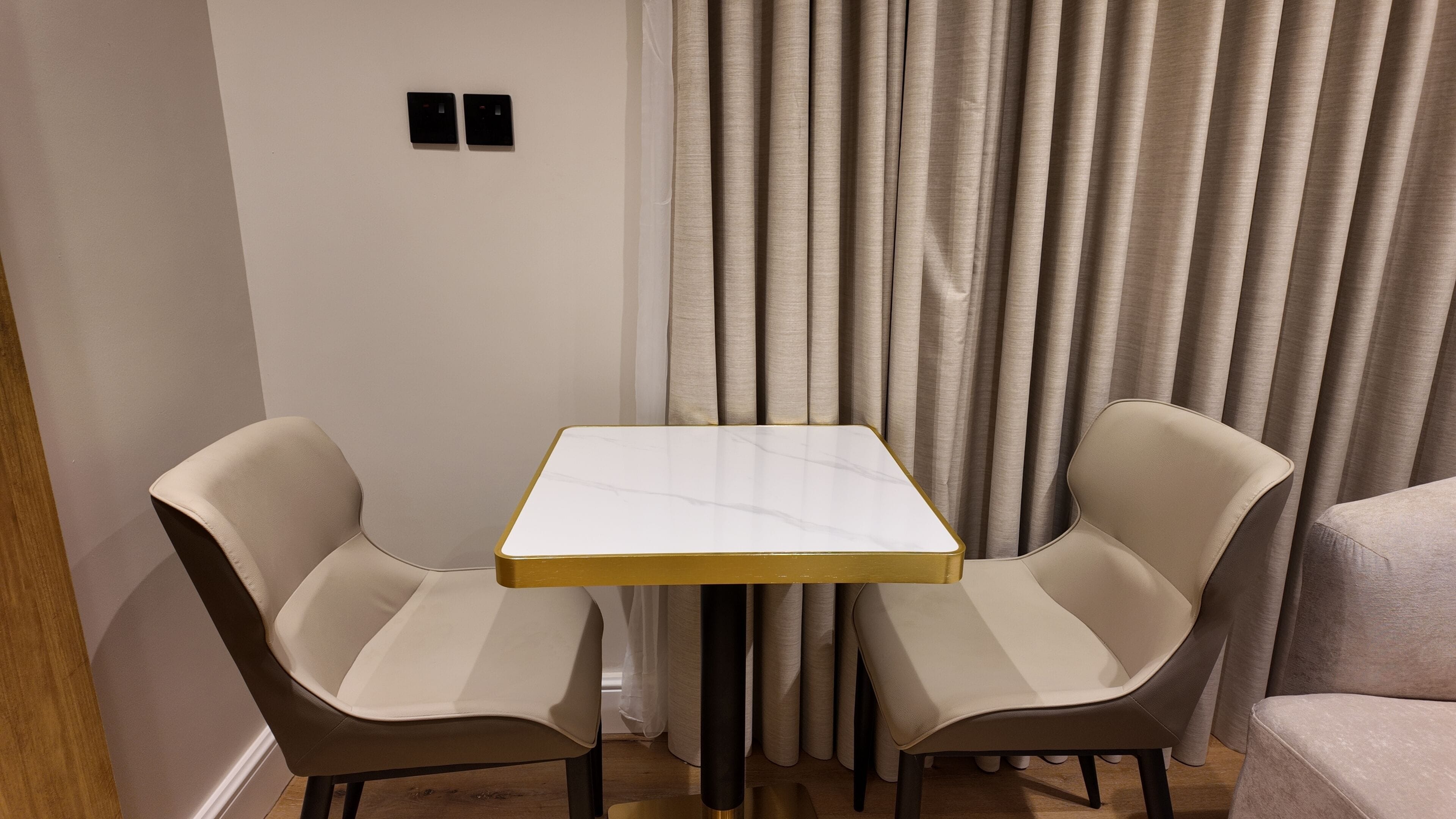 Une petite table carrée, dotée d’un plateau blanc et d’un cadre doré, est flanquée de deux chaises beige aux pieds foncés.