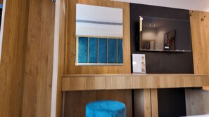 A room with a wooden wall, a window with blue blinds, a flat-screen TV mounted above a wooden cabinet, and a blue ottoman. - AreenPlaza (Jeddah)