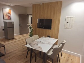 Una zona de menjador amb taula de marbre, quatre cadires, un televisor fixat a una paret de fusta i un frigorífic.