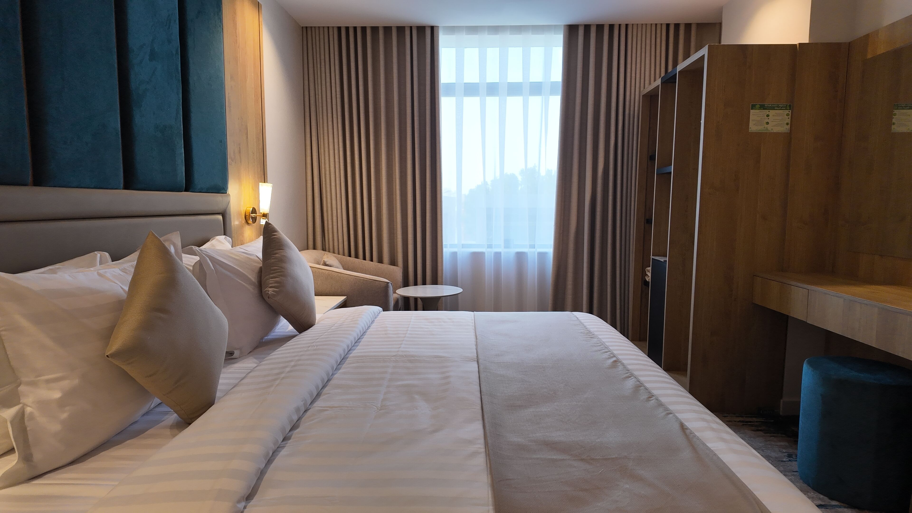 Habitación de hotel con una cama grande, una mesita de noche, una pequeña mesa redonda, un armario y una ventana con cortinas.