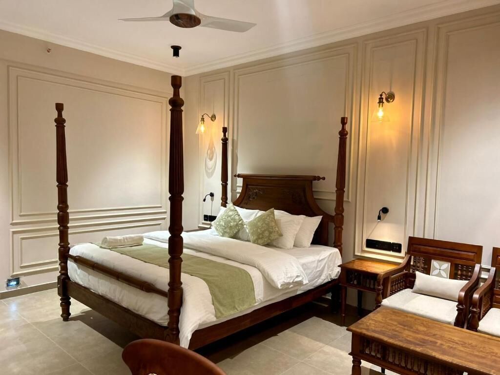 A four-poster bed with a wooden headboard, a bedside table, and a chair in a room with cream-colored walls and a ceiling fan.