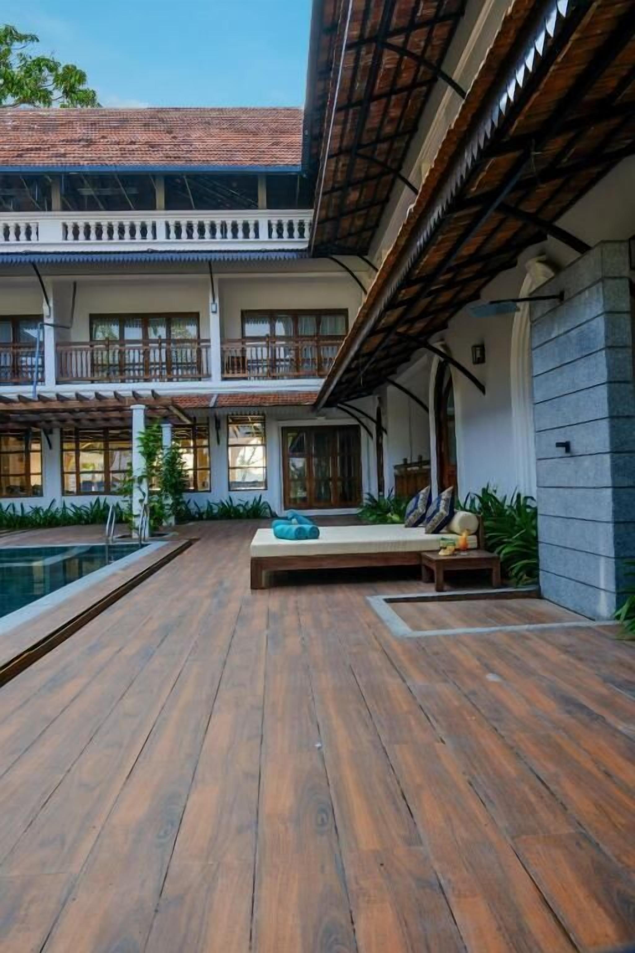 A wooden deck with a pool, a seating area with cushions, and a building with multiple windows and balconies.