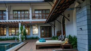 A wooden deck with a pool, a seating area with cushions, and a building with multiple windows and balconies.