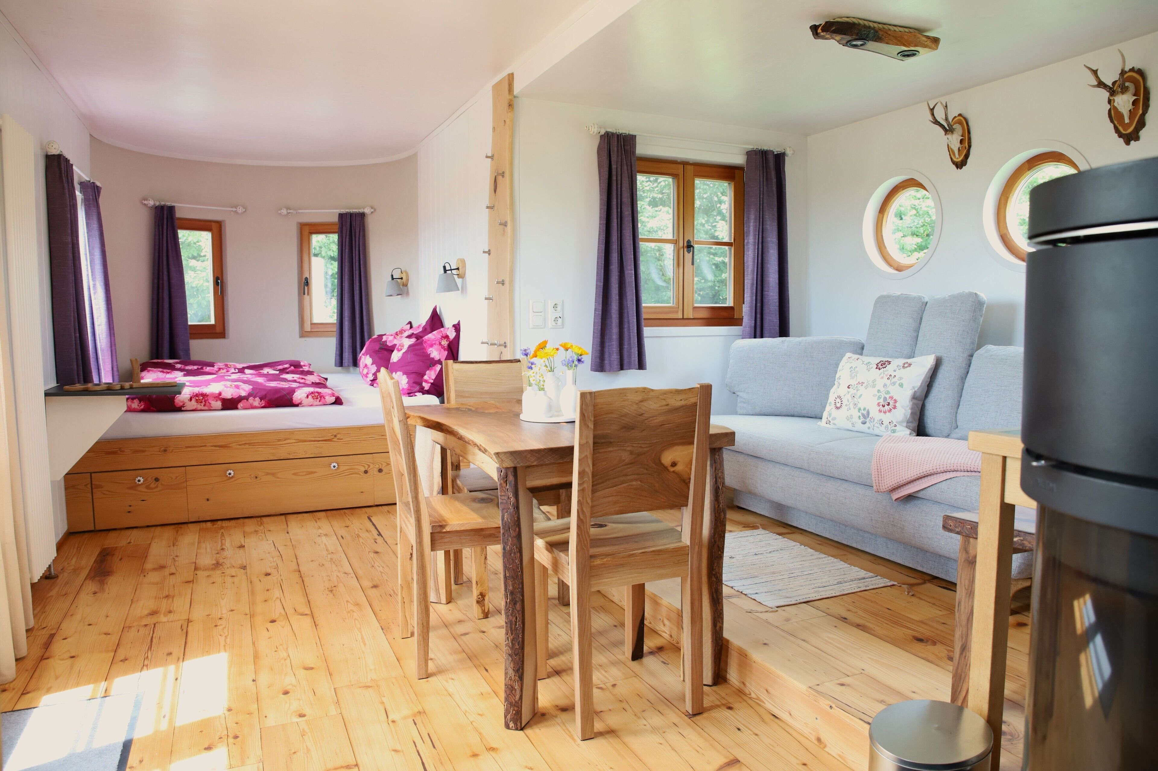 Een gezellige kamer met een houten vloer, een bed met bloemenbeddels, een grijze bank, een eettafel met stoelen en een zwarte koelkast.