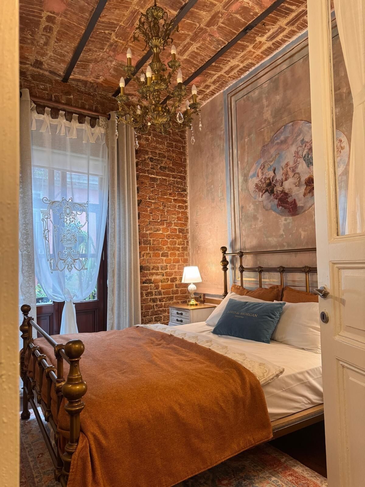 Ein Schlafzimmer mit Backsteinwand, einer Kristallleuchter, einem Bett mit Kopfteil, einem Nachttisch und einem Fenster mit Vorhängen.