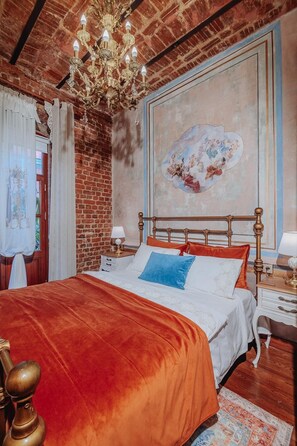A bedroom with a brick wall, a large painting, a chandelier, a bed with a red blanket, and a nightstand with a lamp. - Allegory of Taksim 2B (İstanbul)