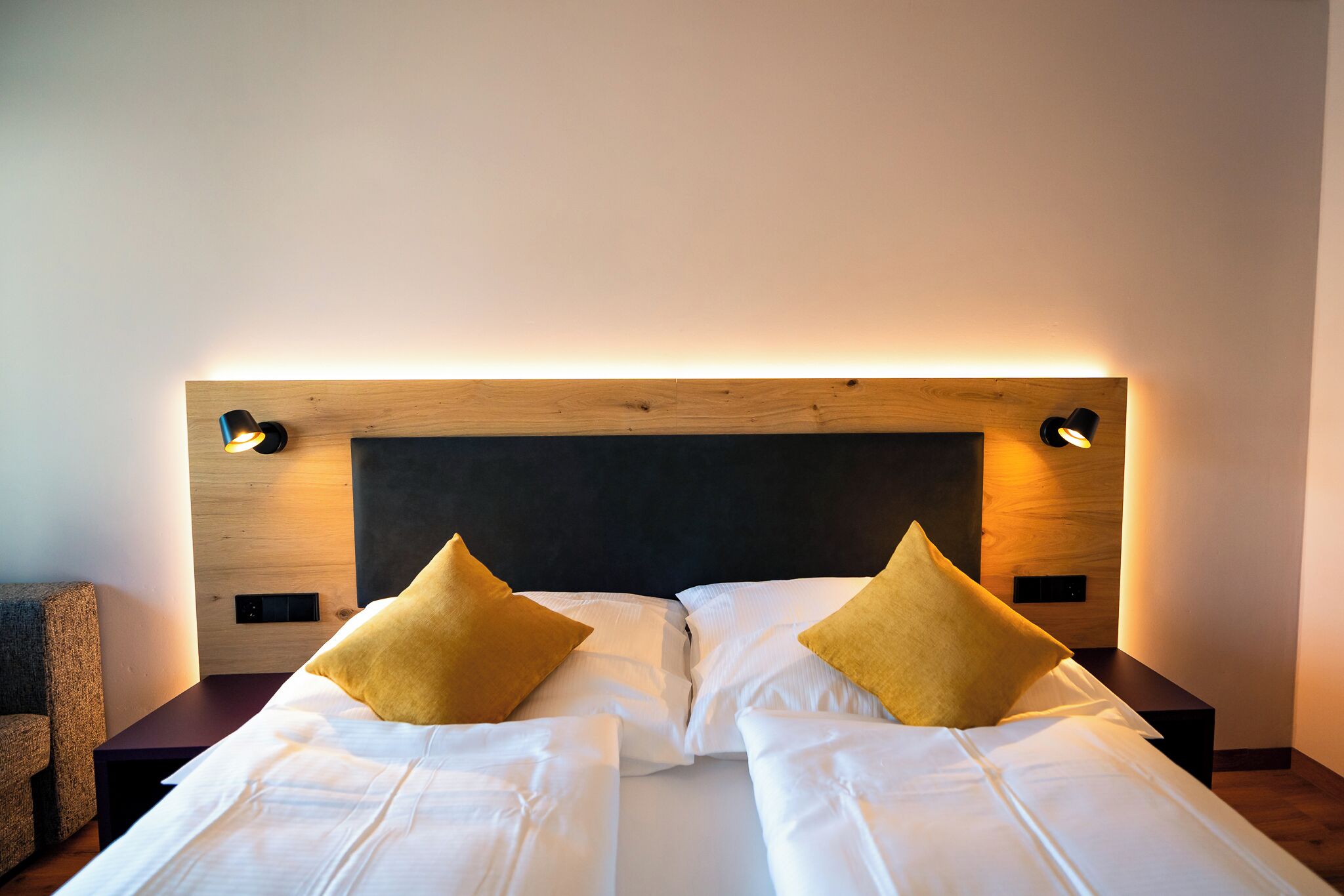 Een tweepersoonsbed met witte lakens en twee mosterdgele kussens, met nachtkastjes met zwarte lampen aan weerszijden, tegen een houten hoofdbord met ingebouwde verlichting.