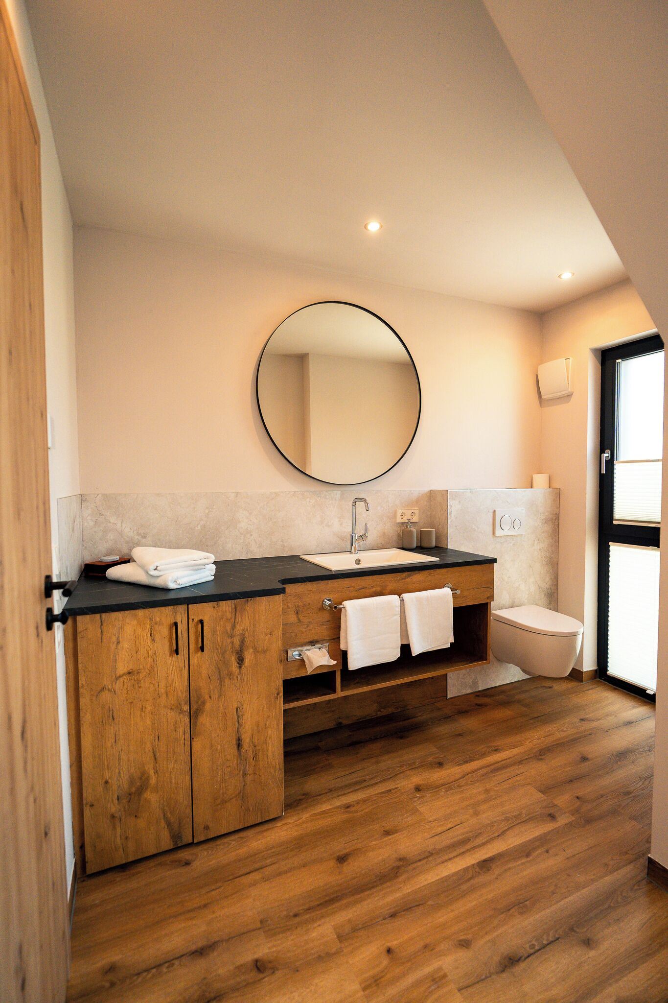 A modern bathroom with a wooden vanity, a round mirror, a sink, and a toilet.