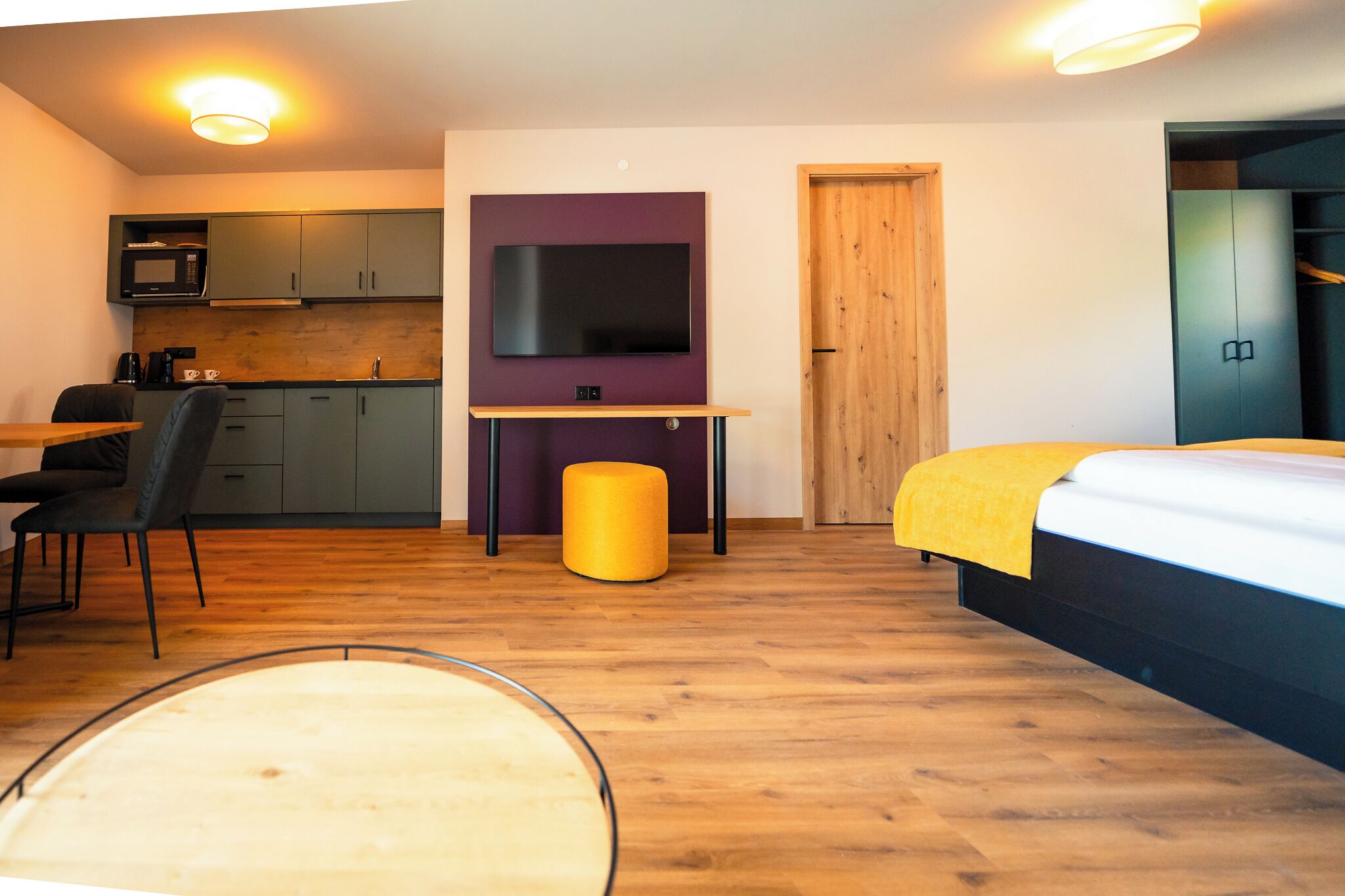 A compact room with a bed, a desk, a chair, a small stool, a television, a wardrobe, and a kitchenette.